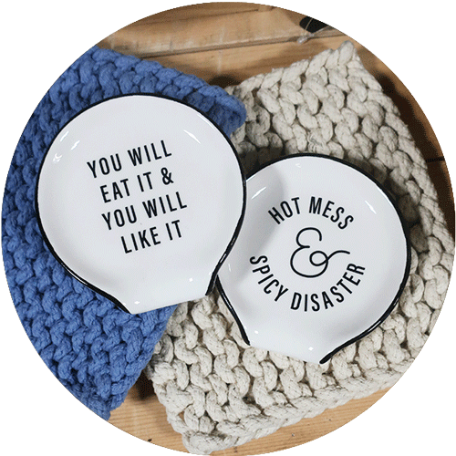 Two spoon rests that say You Will Eat it and You Will Like it, and Hot Mess and Spicy Disaster, on top of blue and cream crochet pot holders.